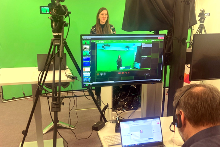 Eine Frau und ein Mann in einem Webcast-Studio. Die Frau steht vor einem Green Screen und spricht in die Kamera, die zweite Person zeichnet auf.