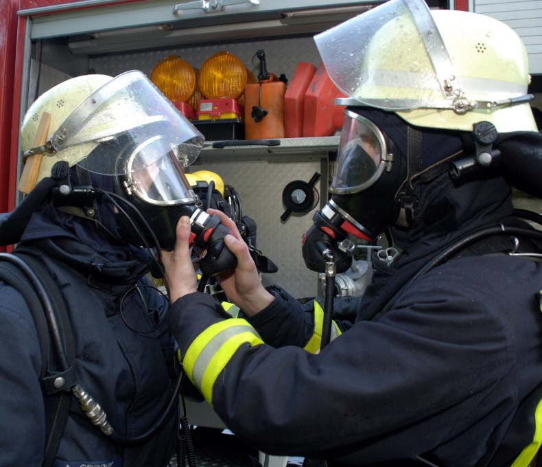 Zwei Feuerwehrleute in vollständiger Schutzausrüstung und Atemschutzmasken stehen vor einem Einsatzfahrzeug. Eine Person hilft der anderen beim Anlegen oder Überprüfen der Atemmaske. Zwei Feuerwehrleute in vollständiger Schutzausrüstung und Atemschutzmasken stehen vor einem Einsatzfahrzeug. Eine Person hilft der anderen beim Anlegen oder Überprüfen der Atemmaske.