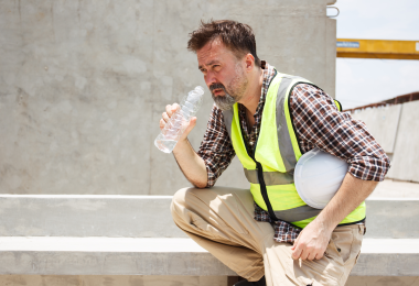 Ein Bauarbeiter mit Warnweste und kariertem Hemd hockt auf einer Baustelle in der Sonne. Er hält einen weißen Schutzhelm unter dem Arm und trinkt aus einer Wasserflasche. Sein Gesicht wirkt angestrengt, die Umgebung besteht aus Betonflächen und Rohbauelementen. (Bild: DG PhotoStock / stock.adobe.com)