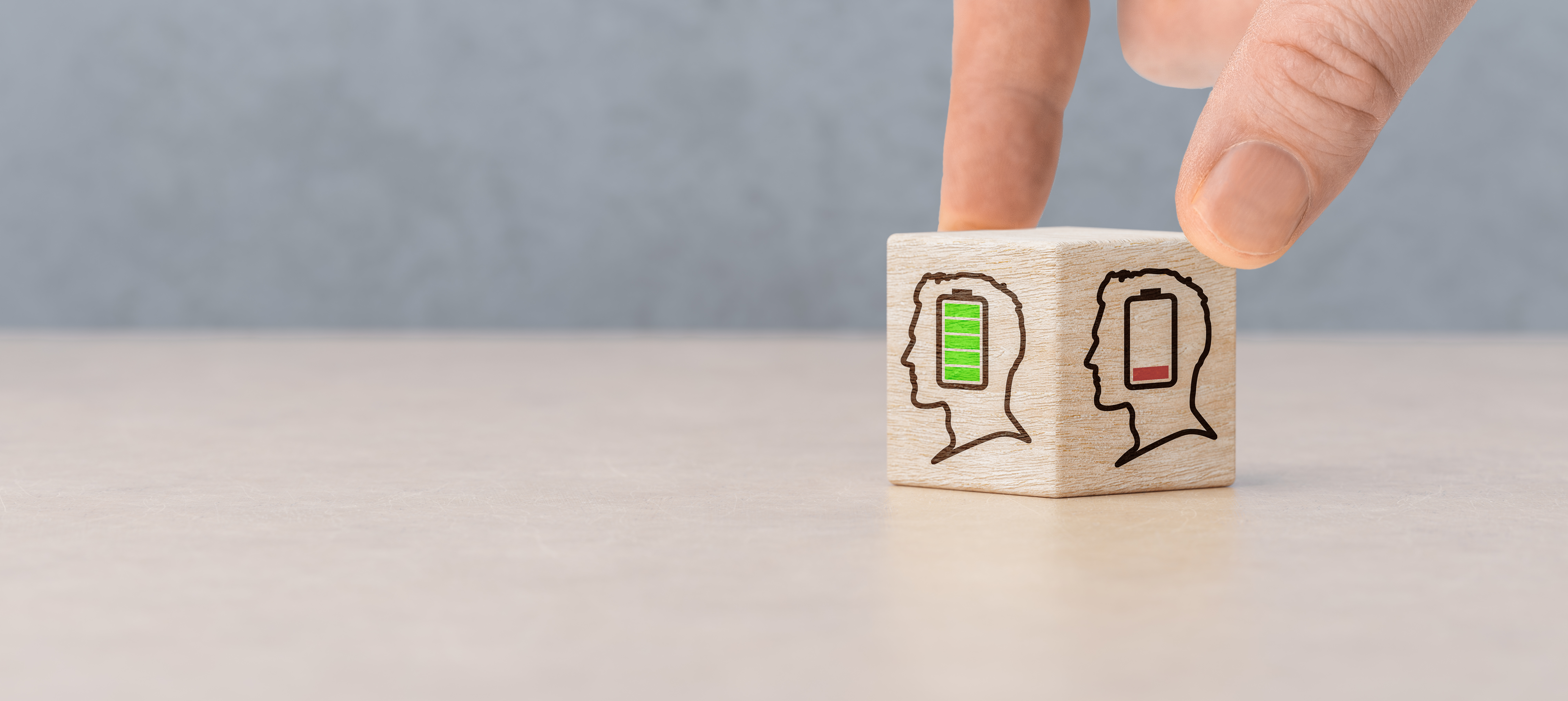 Eine Hand dreht einen kleinen Holzwürfel auf einem Tisch. Auf den sichtbaren Seiten des Würfels sind stilisierte Kopf-Symbole mit Batteriestandsanzeigen abgebildet: einmal mit grün gefüllter Batterie und einmal mit fast leerer roter Batterie. Der Hintergrund ist unscharf und neutral gehalten. (Celt Studio / stock.adobe.com)