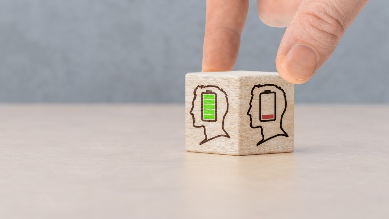 Eine Hand dreht einen kleinen Holzwürfel auf einem Tisch. Auf den sichtbaren Seiten des Würfels sind stilisierte Kopf-Symbole mit Batteriestandsanzeigen abgebildet: einmal mit grün gefüllter Batterie und einmal mit fast leerer roter Batterie. Der Hintergrund ist unscharf und neutral gehalten. (Bild: Celt Studio / stock.adobe.com)