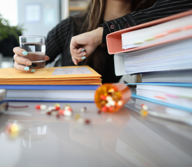 Auf einem Schreibtisch liegen mehrere hohe Stapel von Aktenordnern, Mappen und Dokumenten. Dazwischen sind verstreute bunte Tabletten zu sehen, einige befinden sich noch in einer umgestürzten Medikamentenkapsel. Im Hintergrund hält eine Person ein Glas Wasser in der Hand und stützt sich mit dem Arm auf die Unterlagen. (Bild: H Ko - stock.adobe.com)